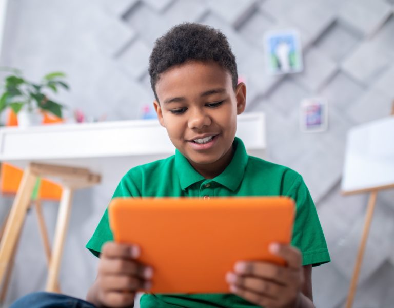 Interesting information. Dark-skinned boy of primary school age in green tshirt looking with interest at orange tablet sitting in room with table and paintings